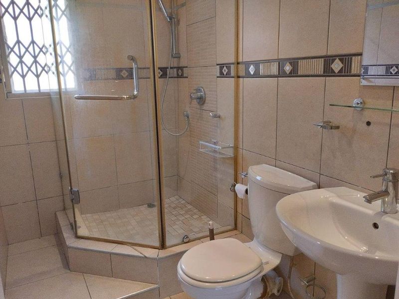 A functional bathroom featuring a corner shower enclosure, pedestal basin, and toilet with neutral wall tiling and a security-gated window.