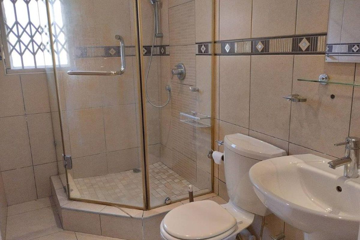 A functional bathroom featuring a corner shower enclosure, pedestal basin, and toilet with neutral wall tiling and a security-gated window.