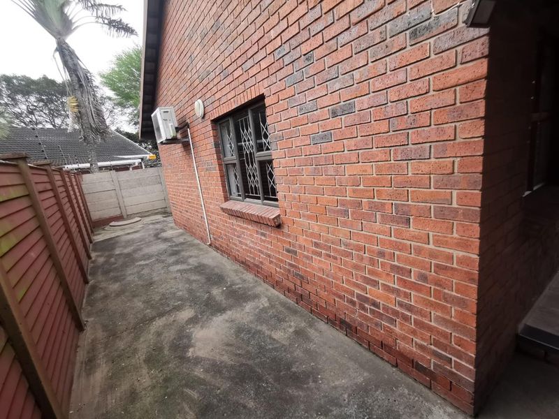 A side view of the apartment showing a brick exterior wall, security-fitted window, external air conditioning unit, and a paved walkway with timber fencing.