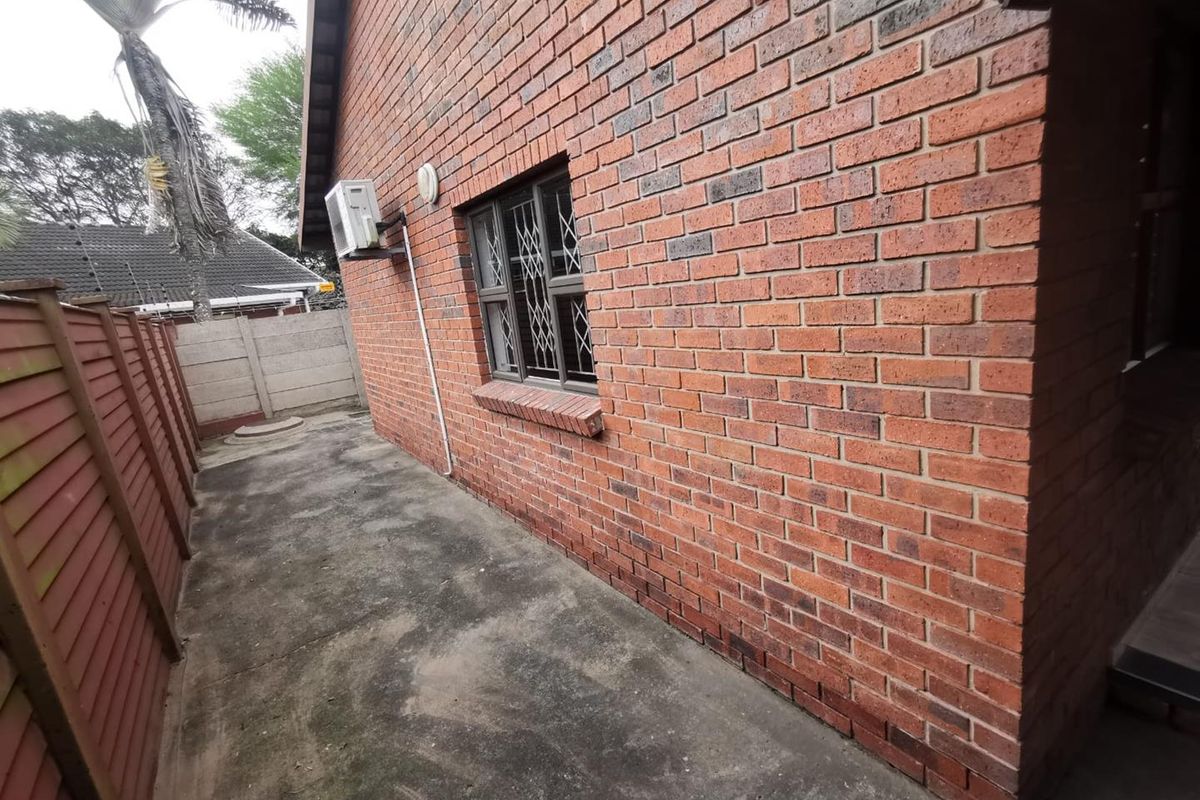 A side view of the apartment showing a brick exterior wall, security-fitted window, external air conditioning unit, and a paved walkway with timber fencing.