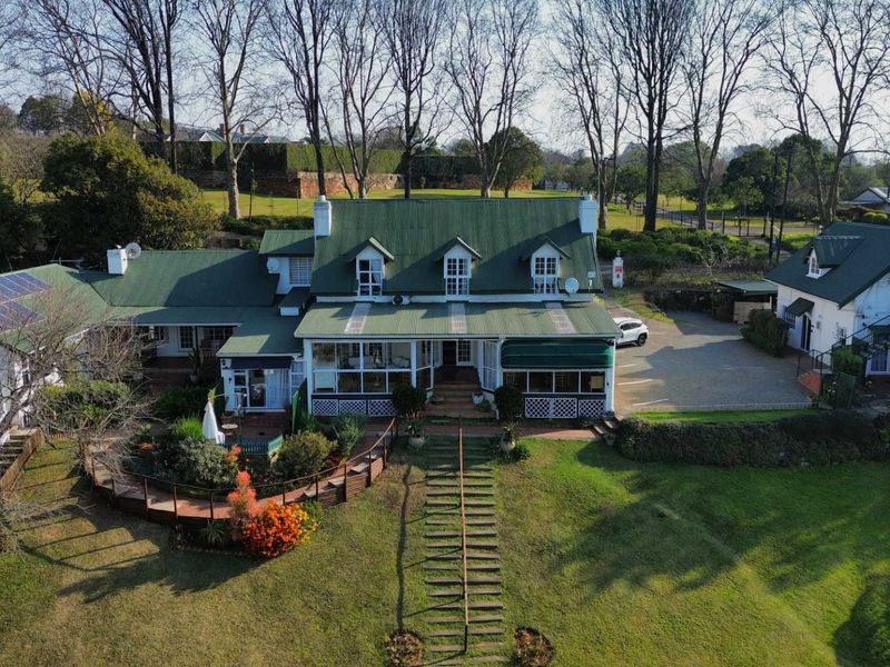 Aerial view of a large property featuring a green metal roof, solar panels, dormer windows, paved driveway, and extensive landscaped grounds.