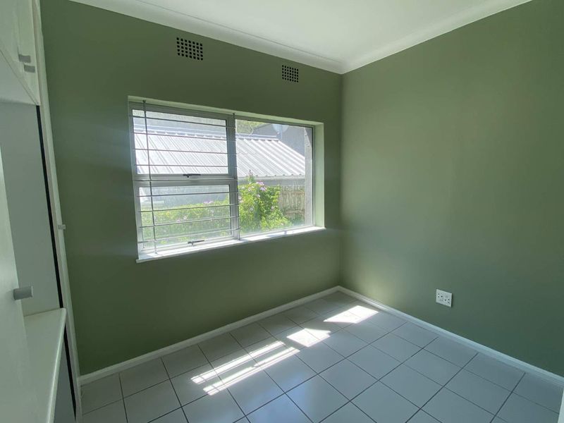 A bright, empty bedroom featuring tiled flooring, built-in storage, and a window with security bars, finished in a neutral green tone.