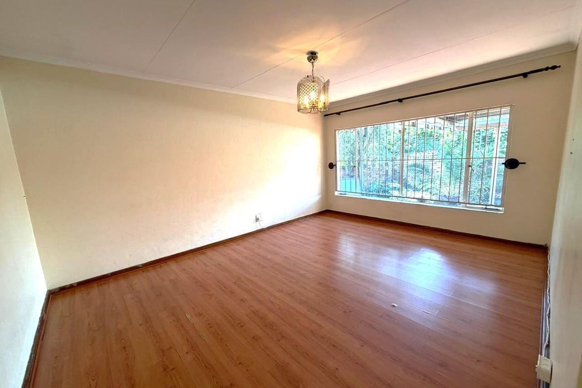 A bright, empty living room featuring laminate wood flooring, a large window with security bars, and a central pendant light fixture.