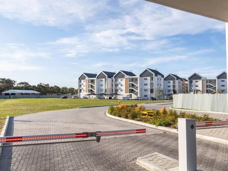 View of the apartment complex entrance featuring a secure boom gate, paved driveway, and modern multi-story residential buildings set in a landscaped estate.