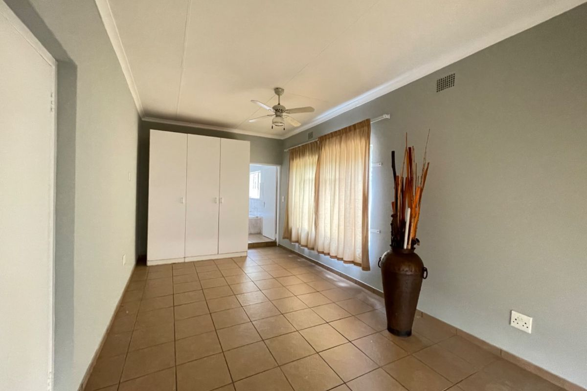 A spacious bedroom featuring neutral wall tones, tiled flooring, built-in cupboards, and a ceiling fan, offering a clean and functional living space.