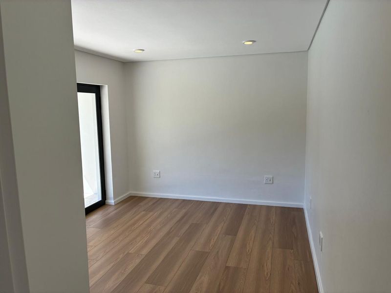 A bright, empty bedroom featuring modern laminate flooring, recessed ceiling lighting, and a black-framed glass door leading to an outdoor area.