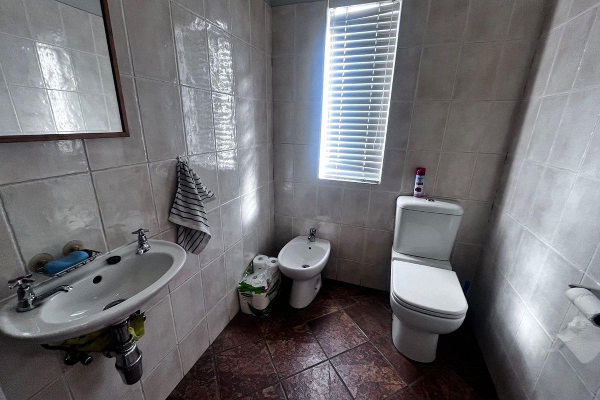 A functional bathroom featuring a wall-mounted basin, bidet, and toilet with full-height wall tiling and a window with fitted blinds.