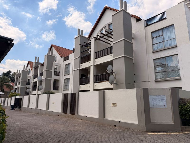 Exterior view of a multi-story apartment complex featuring a paved driveway, perimeter wall, and private balconies with metal railings.