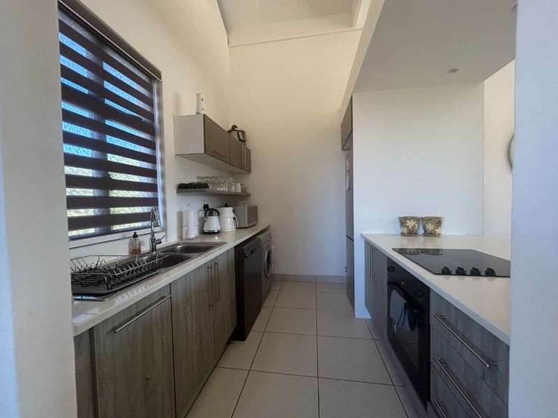 Modern galley-style kitchen featuring wood-grain cabinetry, stone countertops, integrated oven and hob, and tiled flooring with ample natural light.