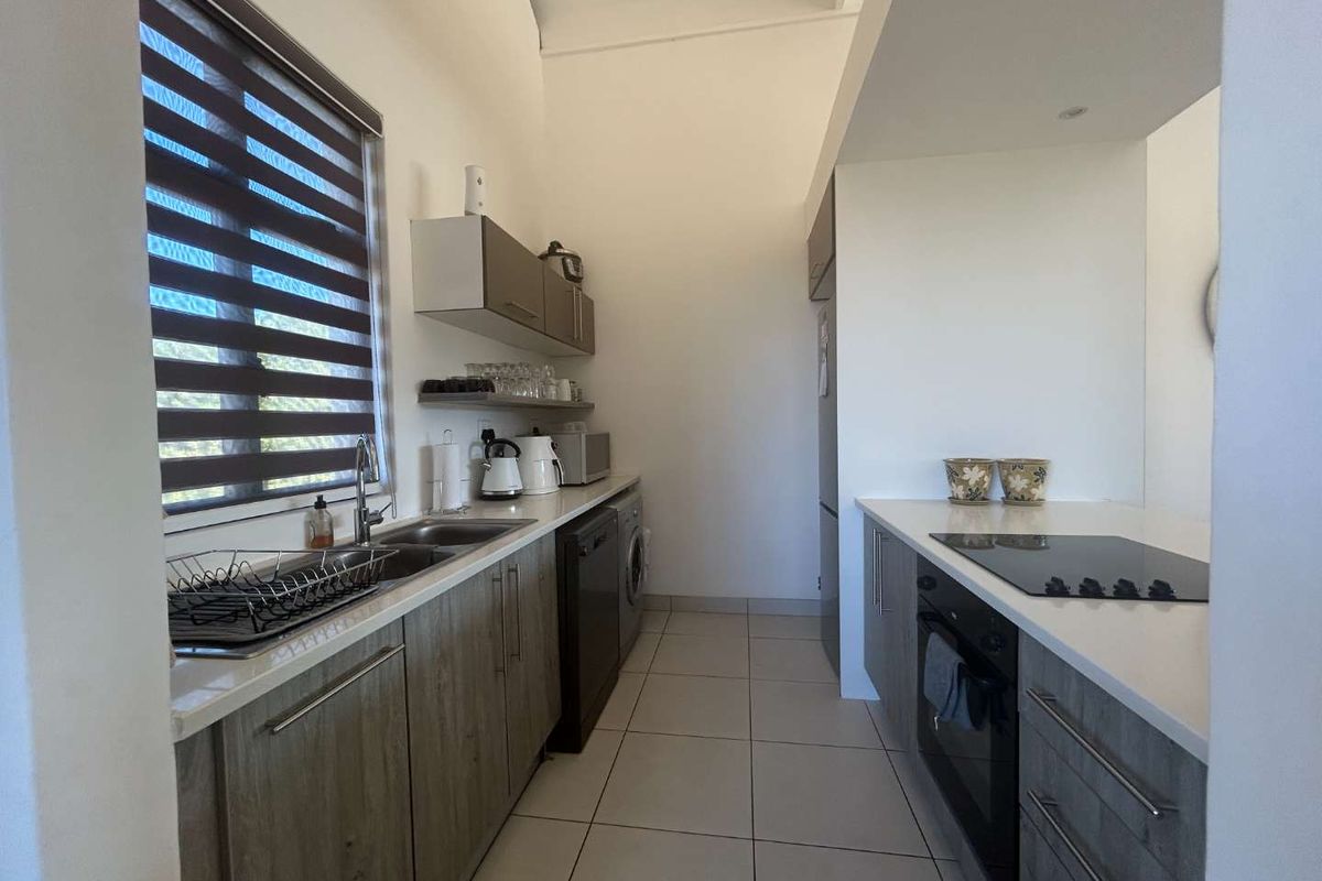 Modern galley-style kitchen featuring wood-grain cabinetry, stone countertops, integrated oven and hob, and tiled flooring with ample natural light.