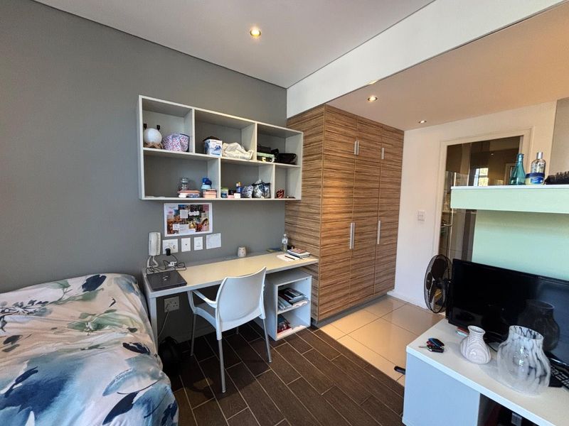 A functional bedroom featuring built-in wooden cabinetry, a wall-mounted desk with shelving, recessed lighting, and tiled flooring.