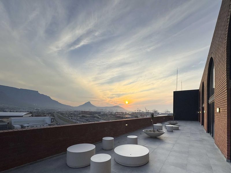 Spacious rooftop balcony featuring modern tiled flooring, brick architectural details, and panoramic sunset views over the city and mountains.
