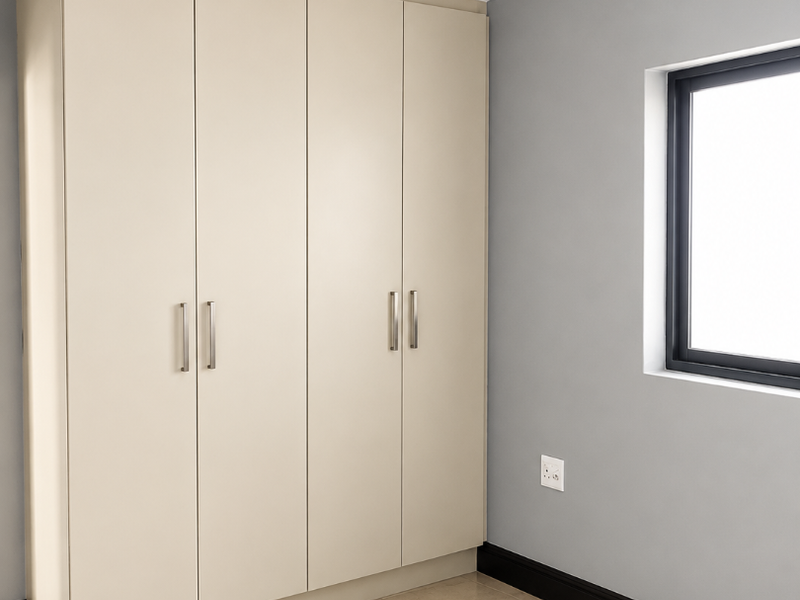 A clean, modern bedroom featuring neutral-toned built-in wardrobes, tiled flooring, and grey walls with contrasting black skirting boards.