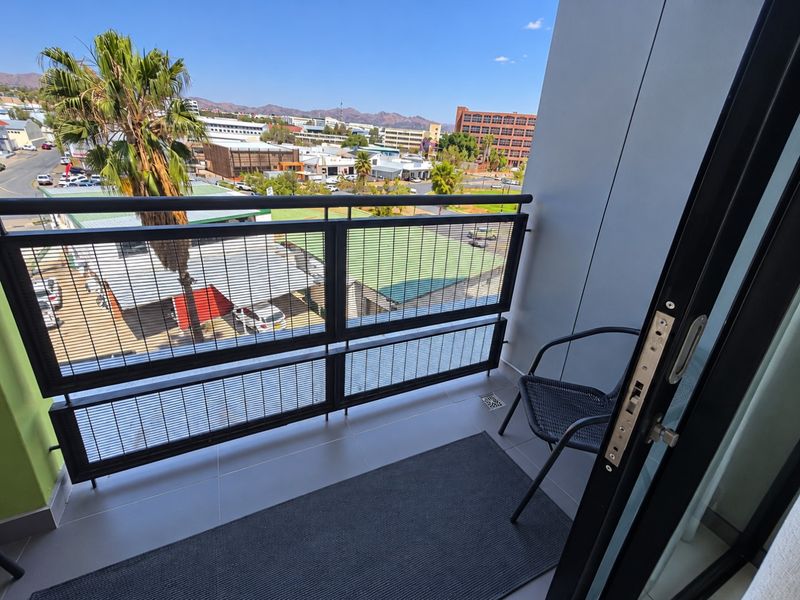A modern balcony featuring tiled flooring, a secure metal balustrade, and a sliding door entrance with views of the surrounding urban area.