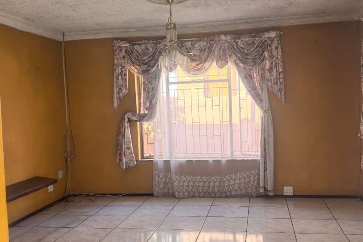An empty living room featuring ceramic floor tiles, a ceiling rose, and security bars on the window. The space requires cosmetic updates.