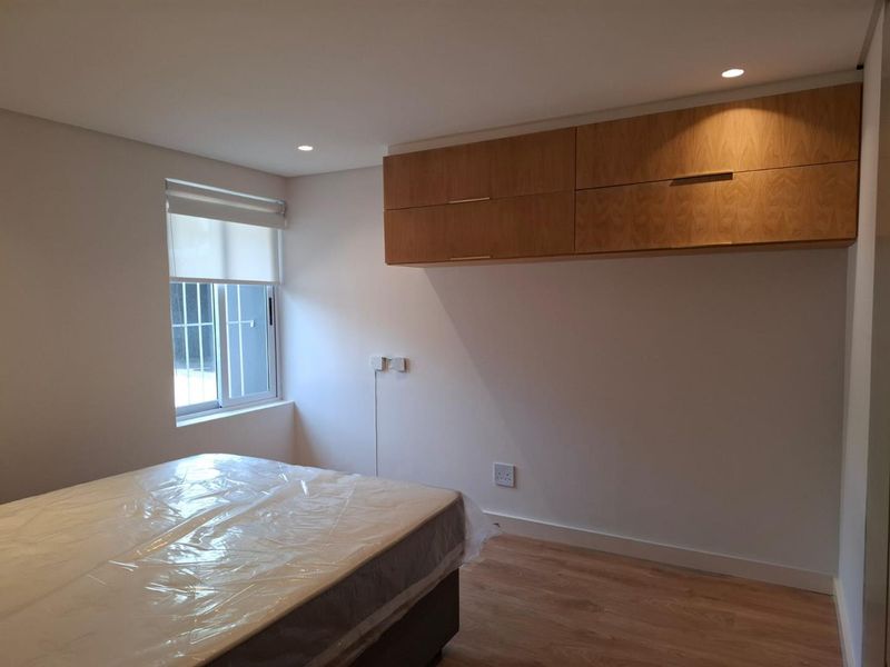 A clean, modern bedroom featuring light wood laminate flooring, built-in overhead cabinetry, recessed lighting, and a window fitted with a roller blind and security bars.