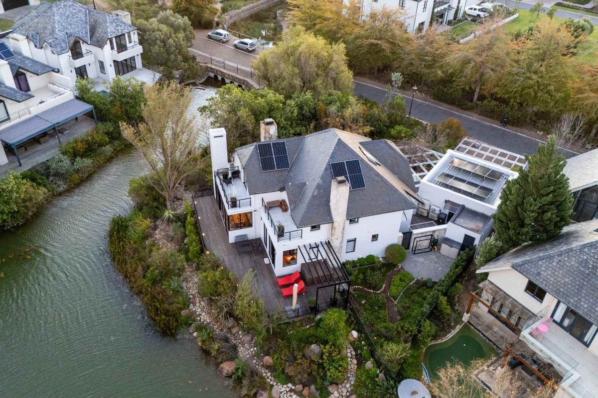 Aerial view of a waterfront residential property featuring a multi-level home with solar panels, a spacious deck, and landscaped garden areas.