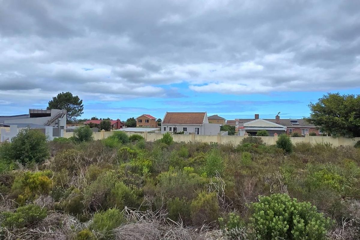 A view of a vacant residential plot featuring natural vegetation, enclosed by a perimeter boundary wall with surrounding neighborhood houses in the background.