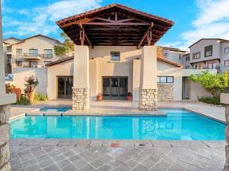 A well-maintained outdoor pool area featuring a covered patio with timber roof structure, stone-clad pillars, and paved surrounds.