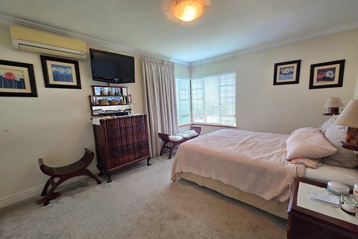 A carpeted bedroom featuring a wall-mounted air conditioning unit, corner windows with blinds, and crown molding.