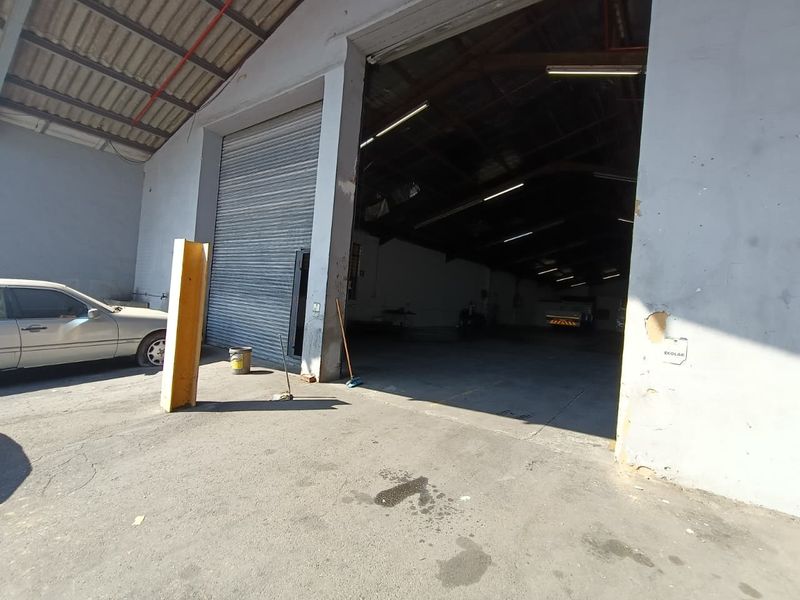 Industrial warehouse exterior featuring a large roller shutter door, concrete apron, and exposed steel roof structure, suitable for storage or manufacturing operations.