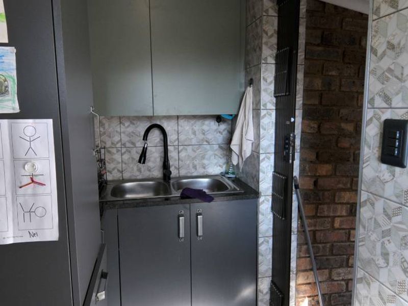 A compact scullery area featuring modern grey cabinetry, a double bowl sink with a gooseneck faucet, and tiled finishes, leading to an exterior doorway.