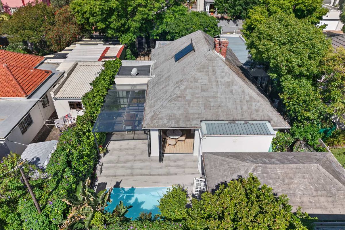 Aerial view of a residential property featuring a swimming pool, a paved patio area with a glass-roofed pergola, and a well-maintained tiled roof.