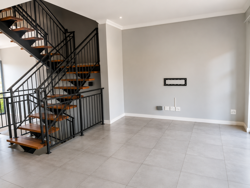 A bright, modern living area featuring a striking open-tread staircase with metal balustrades, neutral tiled flooring, and a wall-mounted TV bracket.