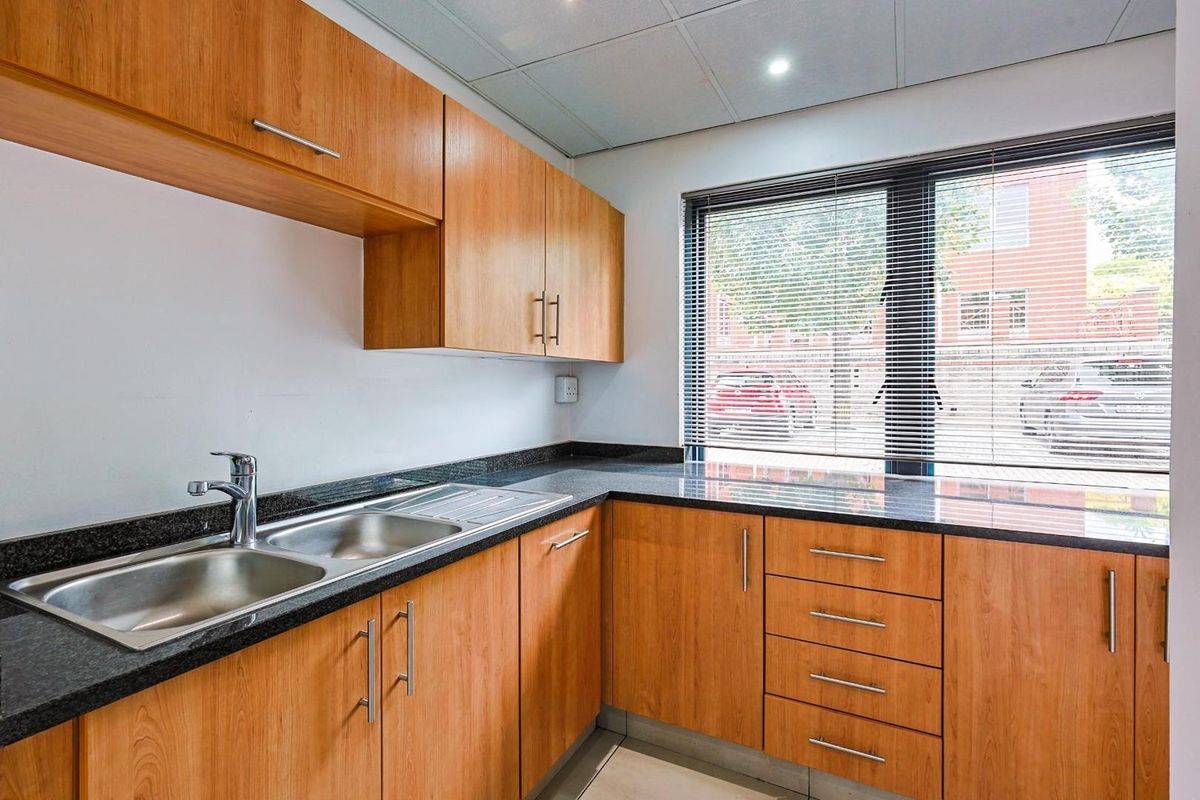 A functional kitchenette featuring wood-finish cabinetry, dark granite countertops, a stainless steel double sink, and a large window with fitted blinds.