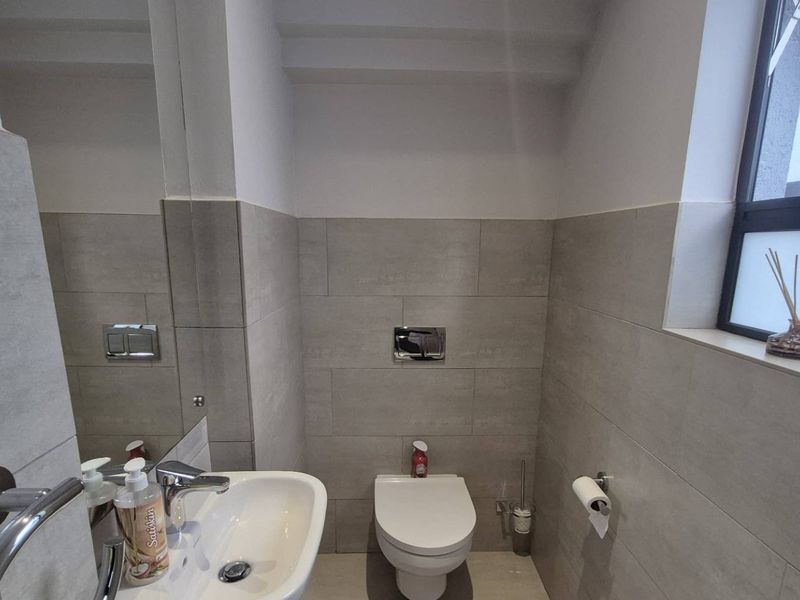 A modern guest bathroom featuring neutral grey wall tiles, a wall-mounted toilet, a sleek basin with a mixer tap, and a frosted window.
