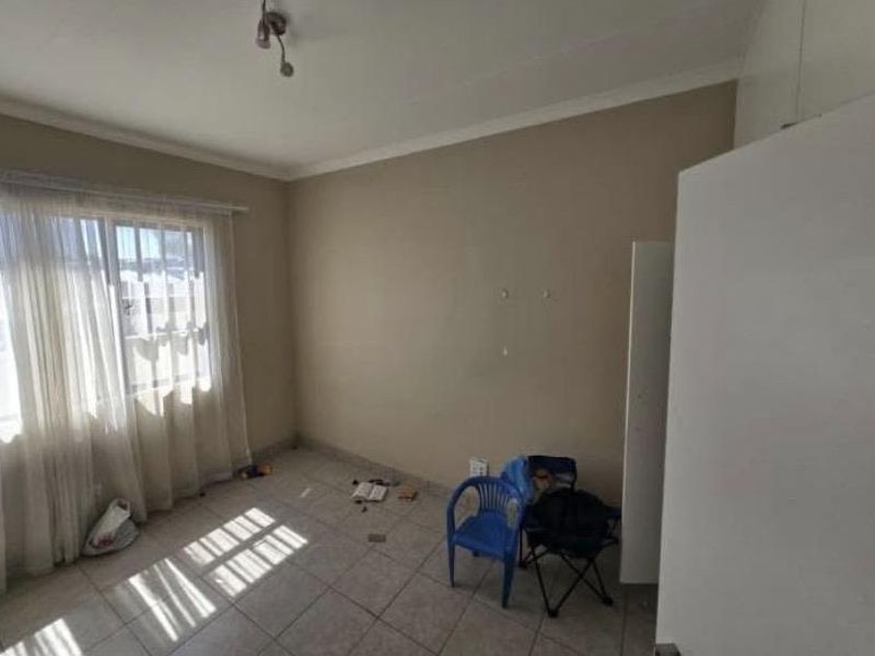 A bright, empty bedroom featuring tiled flooring, built-in cupboards, and a window fitted with security bars.