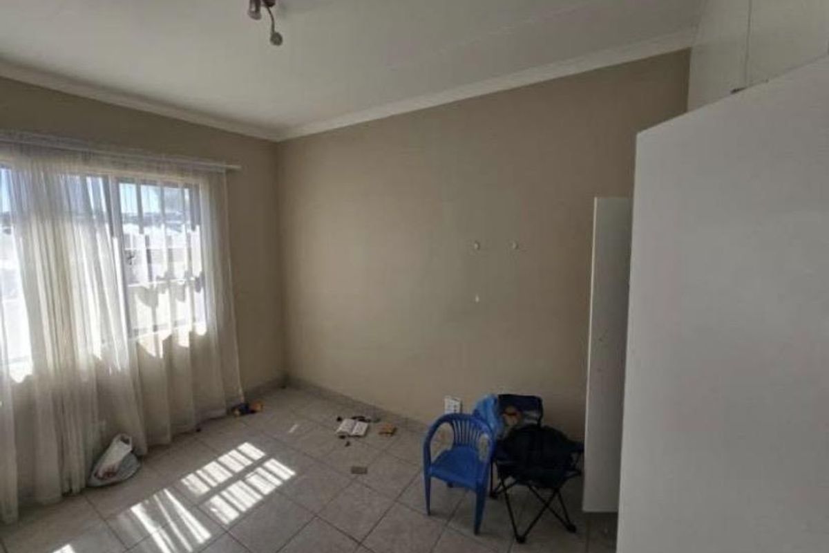A bright, empty bedroom featuring tiled flooring, built-in cupboards, and a window fitted with security bars.