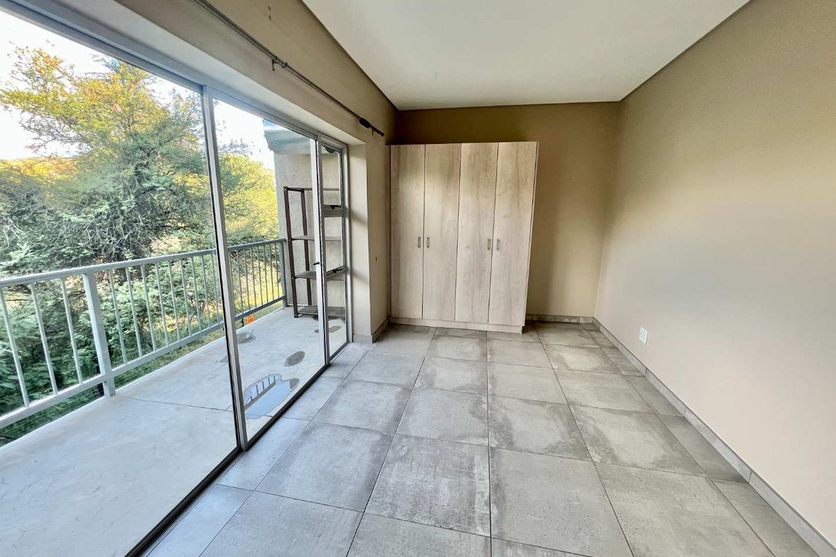 A bright bedroom featuring built-in wooden wardrobes, large sliding glass doors leading to a private balcony, and neutral tiled flooring.