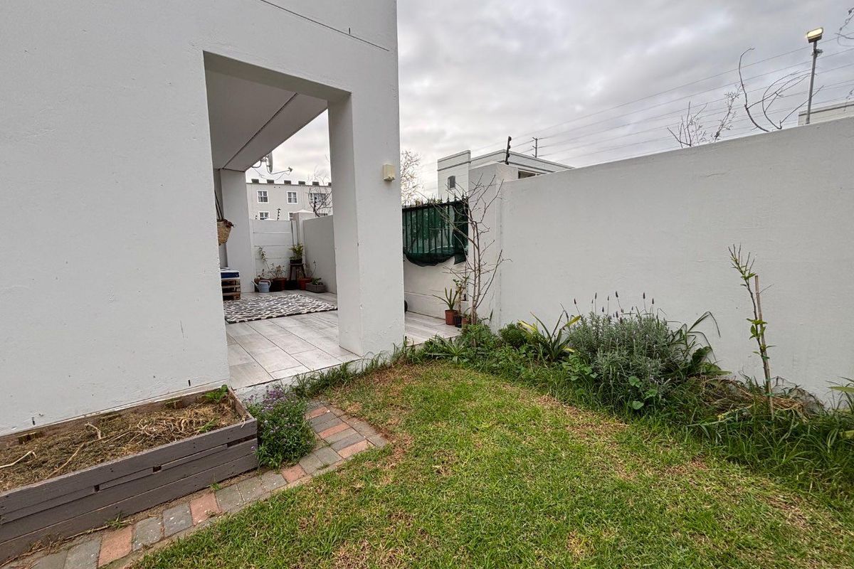 A rear garden area featuring a small lawn, brick-edged garden beds, and a covered patio space with tiled flooring.