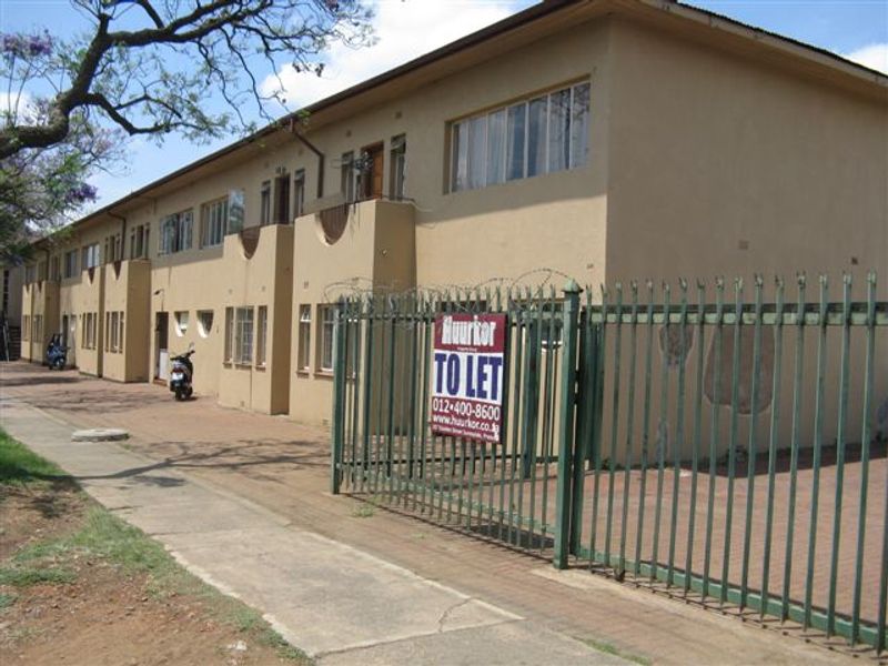 Exterior view of a multi-unit apartment building featuring a paved driveway, security fencing with razor wire, and dated exterior wall finishes.