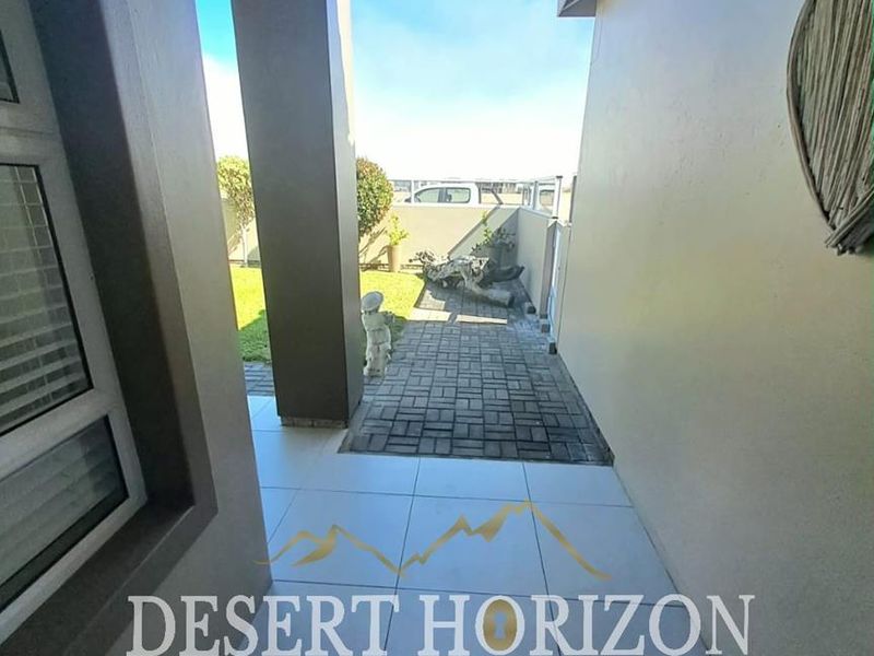 A covered patio area featuring tiled flooring and concrete pillars, leading out to a paved walkway and a small garden space.