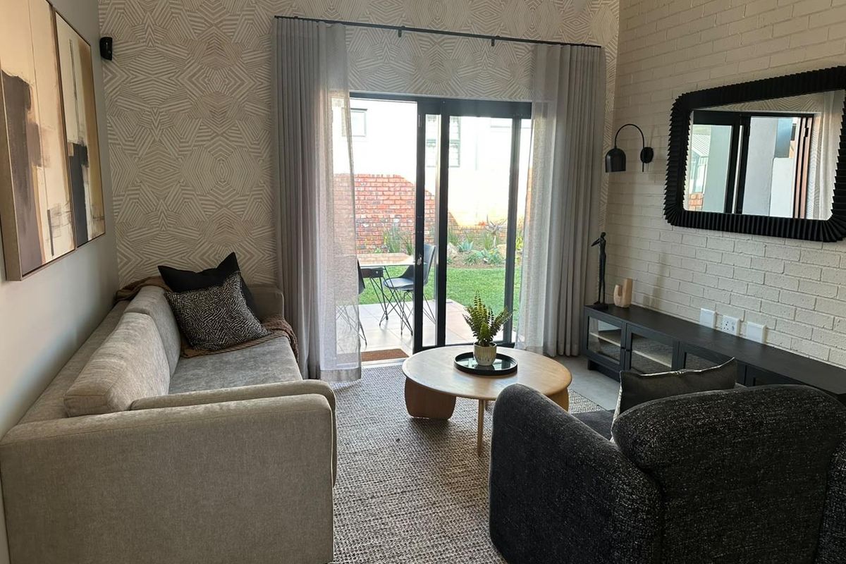 A well-maintained living room featuring a painted brick wall, exposed beam ceiling, and sliding glass doors providing direct access to the rear patio and garden.