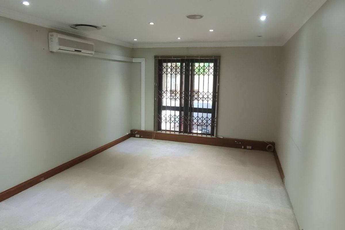 A clean, empty office space featuring neutral walls, carpeted flooring, recessed lighting, and a wall-mounted air conditioning unit with security gate access.