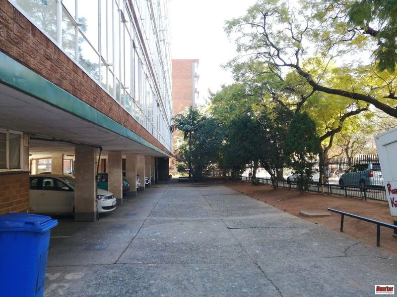 Exterior view of the apartment building featuring a covered parking area, concrete driveway, and established trees along the perimeter fence.