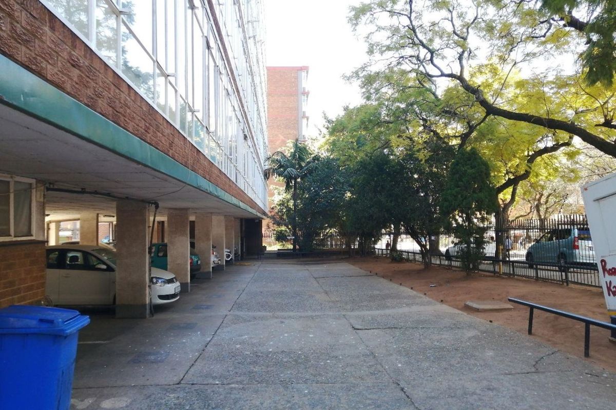 Exterior view of the apartment building featuring a covered parking area, concrete driveway, and established trees along the perimeter fence.