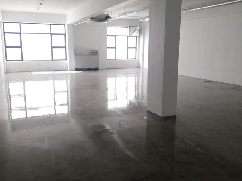 Open-plan office space featuring polished concrete flooring, exposed ceiling services, ducted air conditioning, and a small kitchenette area with large windows.