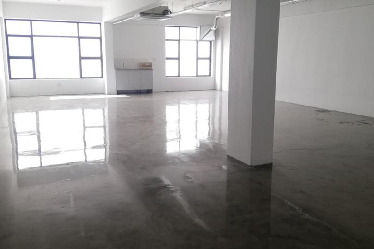 Open-plan office space featuring polished concrete flooring, exposed ceiling services, ducted air conditioning, and a small kitchenette area with large windows.