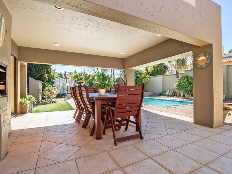 A spacious covered patio featuring tiled flooring, recessed lighting, and a built-in braai, overlooking the garden and swimming pool area.