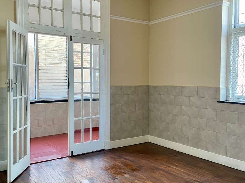 An empty living room featuring original hardwood flooring, tiled wainscoting, and classic French doors leading to an enclosed balcony area.