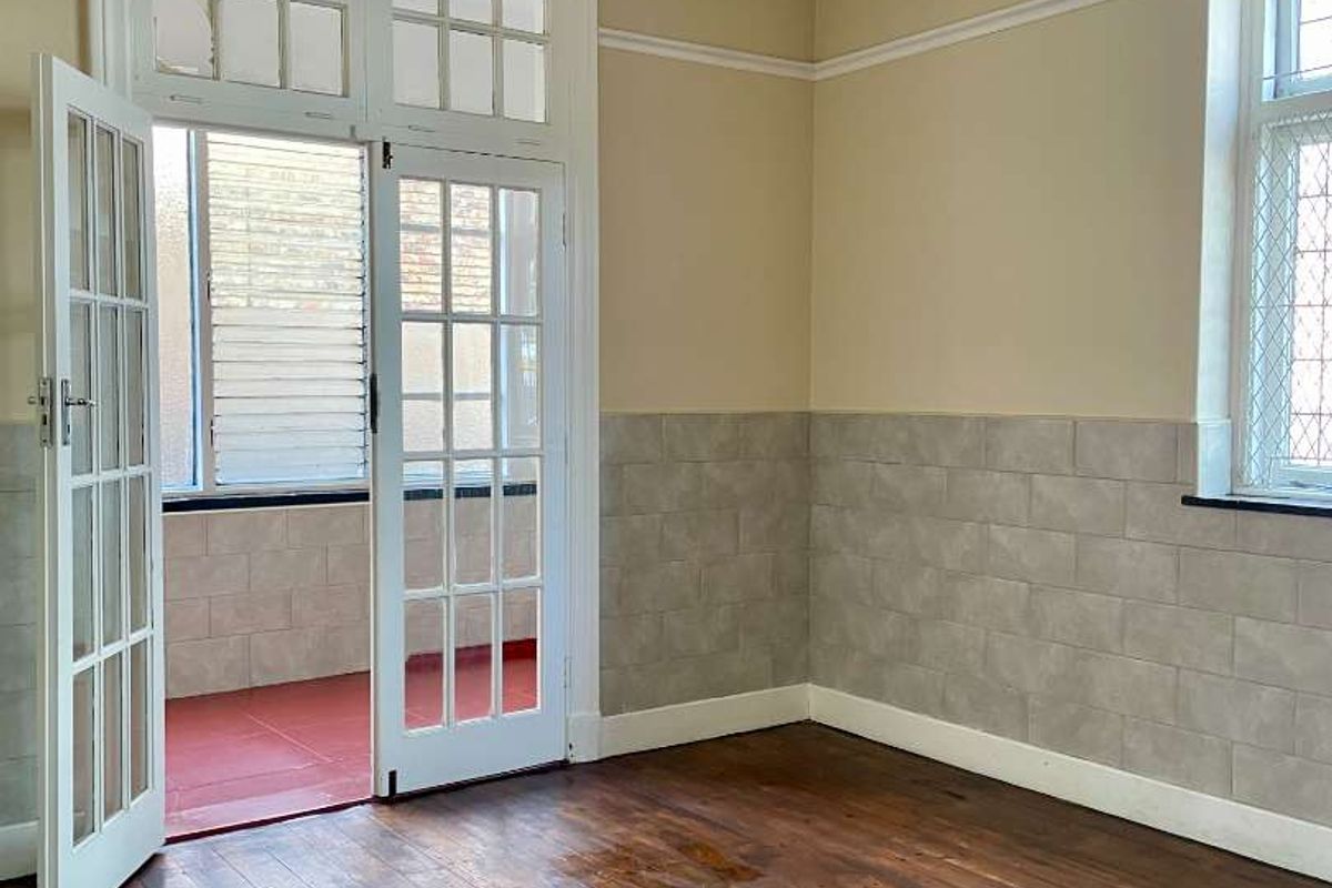 An empty living room featuring original hardwood flooring, tiled wainscoting, and classic French doors leading to an enclosed balcony area.