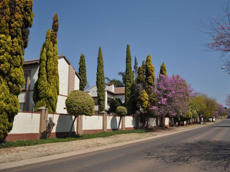 A street-level view of the apartment complex perimeter, featuring a well-maintained boundary wall and mature landscaping along the roadside.
