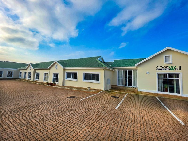 Front exterior view of the CareWell Stilbaai retirement facility, featuring a paved parking area and well-maintained building facade with green metal roofing.