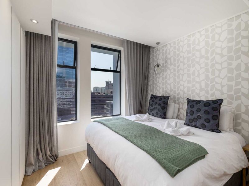 A modern, well-maintained bedroom featuring light laminate flooring, built-in wardrobes, and large windows offering an urban city view.