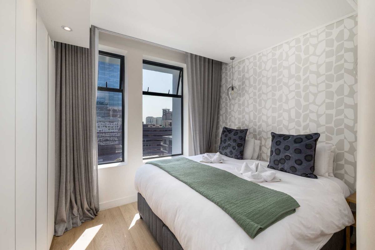 A modern, well-maintained bedroom featuring light laminate flooring, built-in wardrobes, and large windows offering an urban city view.