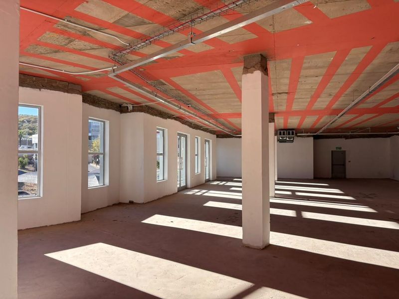 An open-plan office space in shell condition, featuring exposed concrete ceilings, structural columns, and large windows providing ample natural light.
